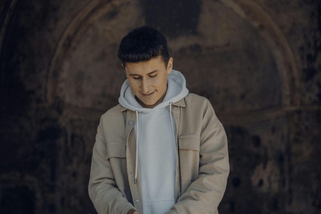 A young man in a beige jacket smiles thoughtfully while looking down, creating a warm, stylish portrait.