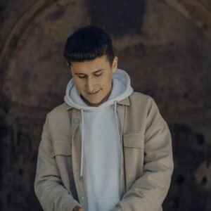 A young man in a beige jacket smiles thoughtfully while looking down, creating a warm, stylish portrait.